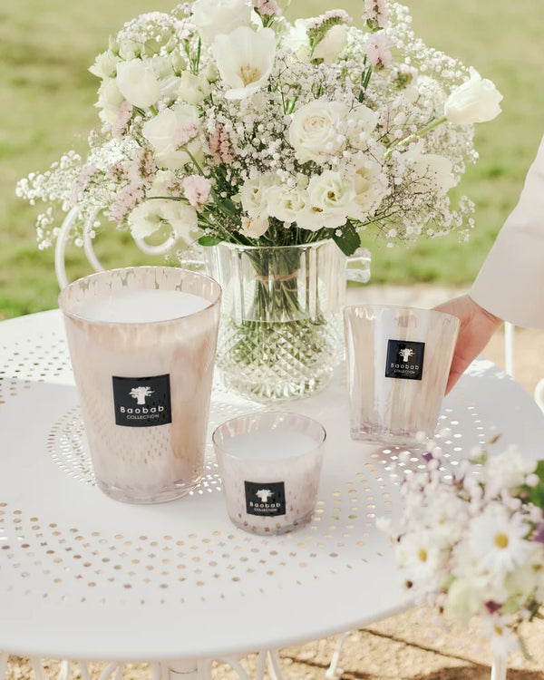 Three scented Pearls Candle - White glass candles in varying sizes arranged on a white metal table with a vase of white flowers and scattered white pearls.