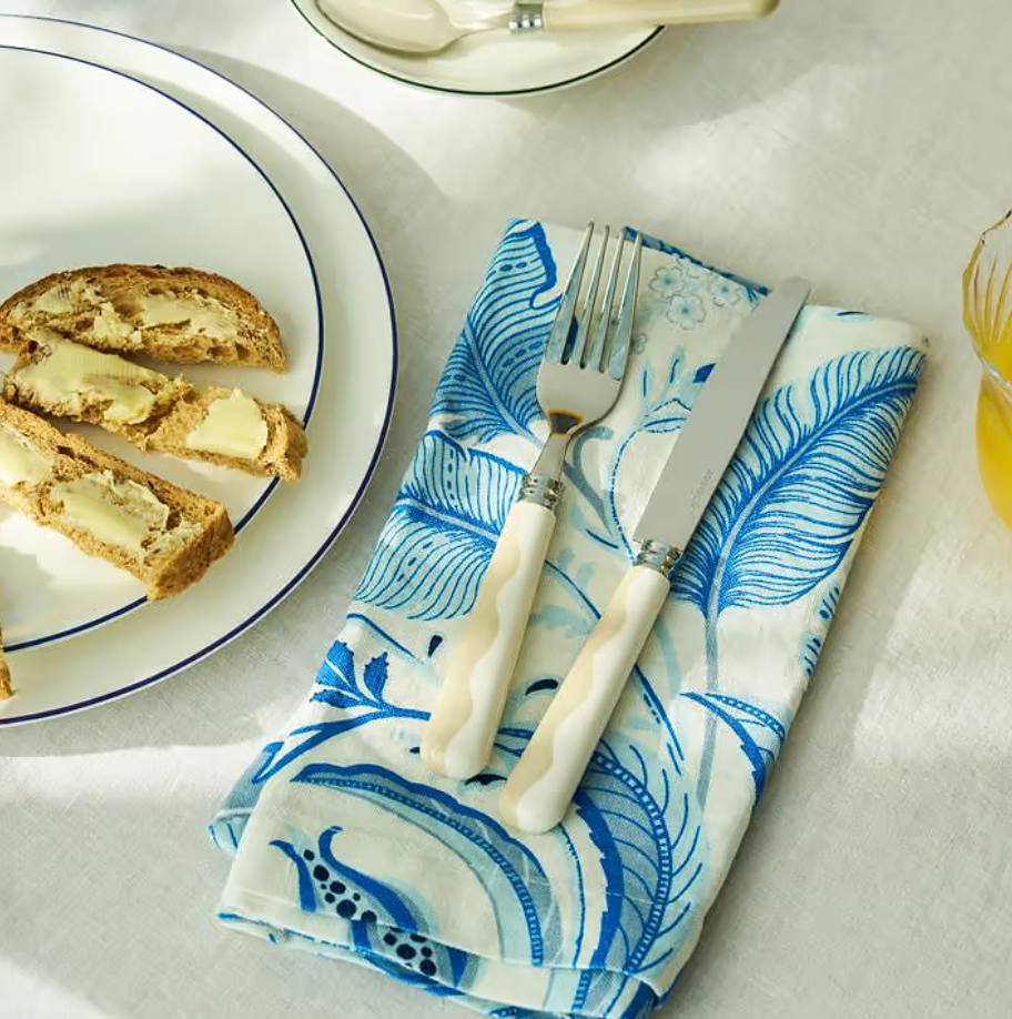 A plate with bread and butter sits on a white tablecloth next to a fork and knife from the Addison Ross Wave Outdoor Flatware 4 Sets (Service for 4), resting on a blue and white patterned napkin, with a glass of orange juice nearby.