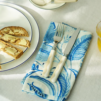 A plate with bread and butter sits on a white tablecloth next to a fork and knife from the Addison Ross Wave Outdoor Flatware 4 Sets (Service for 4), resting on a blue and white patterned napkin, with a glass of orange juice nearby.