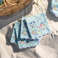The Day At The Beach Cocktail Napkin Set of 4 by Pomegranate features light blue scalloped napkins embroidered with beach scenes, arranged fan-like on sand next to a woven basket for a vibrant touch to your table decor.