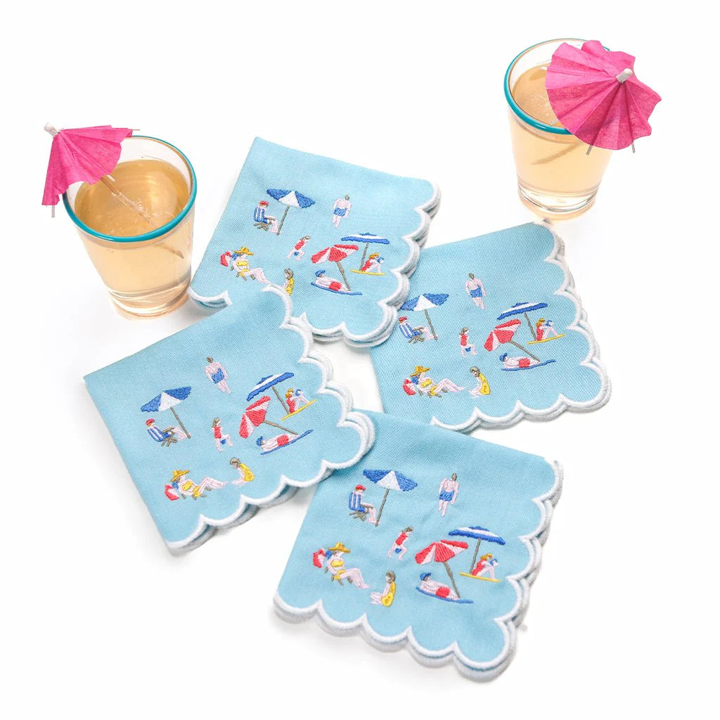 Two drinks with pink paper umbrellas sit beside four Day At The Beach Cocktail Napkins by Pomegranate, each decorated with vibrant beach scenes—making a perfect set of festive drink accessories and colorful table décor.