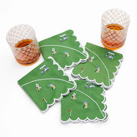 Two glasses rest beside the Day At The Golf Course Cocktail Napkin Set of 4 by Pomegranate—green, scalloped-edge napkins featuring golfers, golf carts, and flags—to add playful charm to your tableware on a crisp white background.