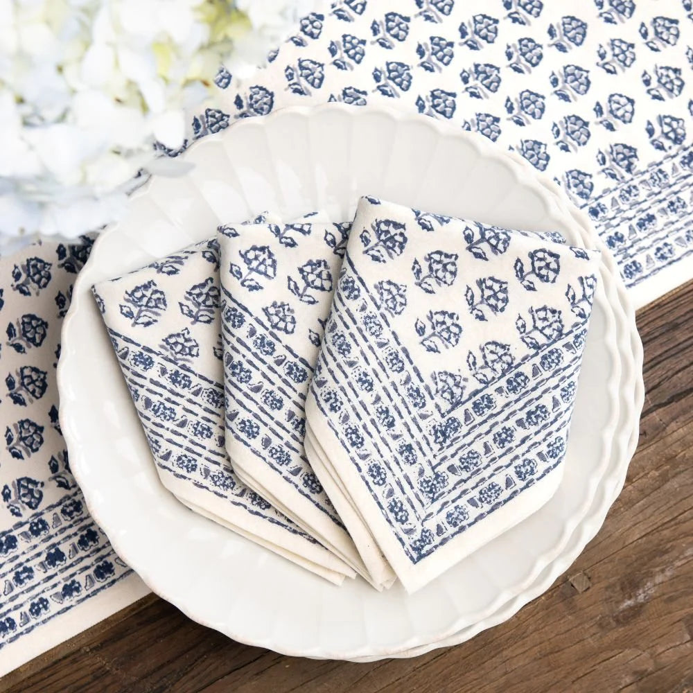 The Elise Napkin Set of 4 - Blue by Pomegranate is displayed neatly folded on a white scalloped plate atop a wooden table, with a matching blue and white runner and white floral decor in the background.