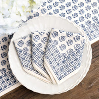 The Elise Napkin Set of 4 - Blue by Pomegranate is displayed neatly folded on a white scalloped plate atop a wooden table, with a matching blue and white runner and white floral decor in the background.