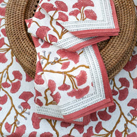 A close-up of the Ginkgo Napkin Set of 4 - Coral by Pomegranate, featuring folded napkins with pink and gold ginkgo motifs in a wicker basket on a matching floral tablecloth—ideal for elegant table decor.