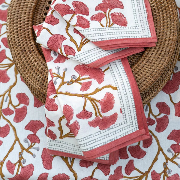 A close-up of the Ginkgo Napkin Set of 4 - Coral by Pomegranate, featuring folded napkins with pink and gold ginkgo motifs in a wicker basket on a matching floral tablecloth—ideal for elegant table decor.