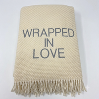 The Wrapped in Love Throw by A Soft Idea is a super soft, cream-colored woven blanket with fringed edges and "WRAPPED IN LOVE" embroidered in gray—an ideal holiday gift.