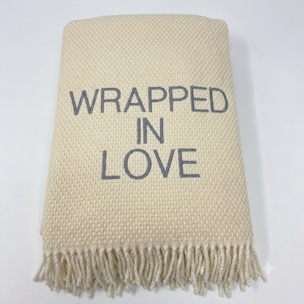 The Wrapped in Love Throw by A Soft Idea is a super soft, cream-colored woven blanket with fringed edges and "WRAPPED IN LOVE" embroidered in gray—an ideal holiday gift.