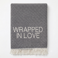A Soft Idea’s Wrapped in Love Throw is a folded dark gray woven blanket with white fringed edges. Super soft, machine washable, and “WRAPPED IN LOVE” stitched in white—perfect for gifting.