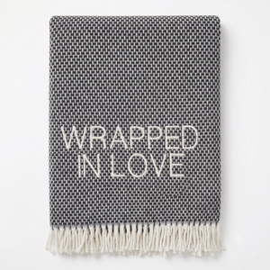 A Soft Idea’s Wrapped in Love Throw is a folded dark gray woven blanket with white fringed edges. Super soft, machine washable, and “WRAPPED IN LOVE” stitched in white—perfect for gifting.