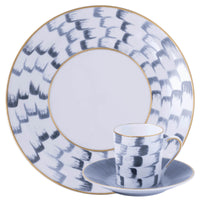 A white Marie Daâge Panache porcelain dinner plate, cup, and saucer, all decorated with a pattern of dark grey brushstrokes and gold rims.