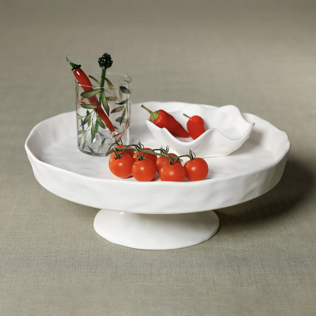 The Zodax Alto Ceramic Pedestal Bowl displays cherry tomatoes and a small dish of red chili peppers. Next to it, a glass with green leaf design contains more chilies, all set against a simple neutral background.