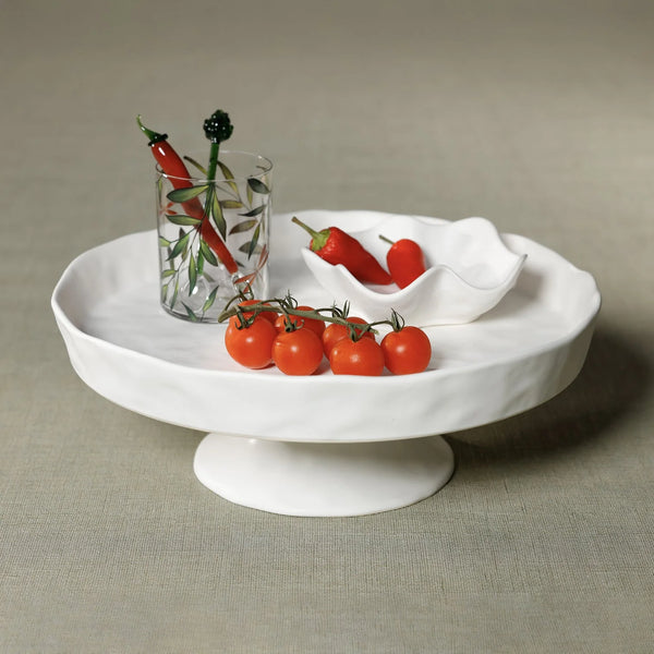 The Zodax Alto Ceramic Pedestal Bowl displays cherry tomatoes and a small dish of red chili peppers. Next to it, a glass with green leaf design contains more chilies, all set against a simple neutral background.