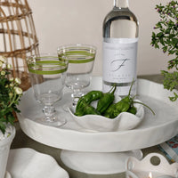 A white platter displays two green-striped glasses, a bottle of sparkling water, and the Alto Ceramic Condiment Bowl by Zodax filled with green peppers, all arranged with potted plants and a lit candle on modern tableware.