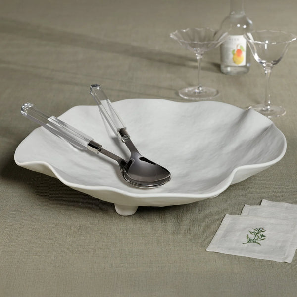 The Zodax Alto Ceramic Footed Bowl, paired with metal and clear plastic tongs, sits on a taupe tablecloth. Behind it are two martini glasses, a bottle featuring a pear label, and napkins embroidered with green accents.