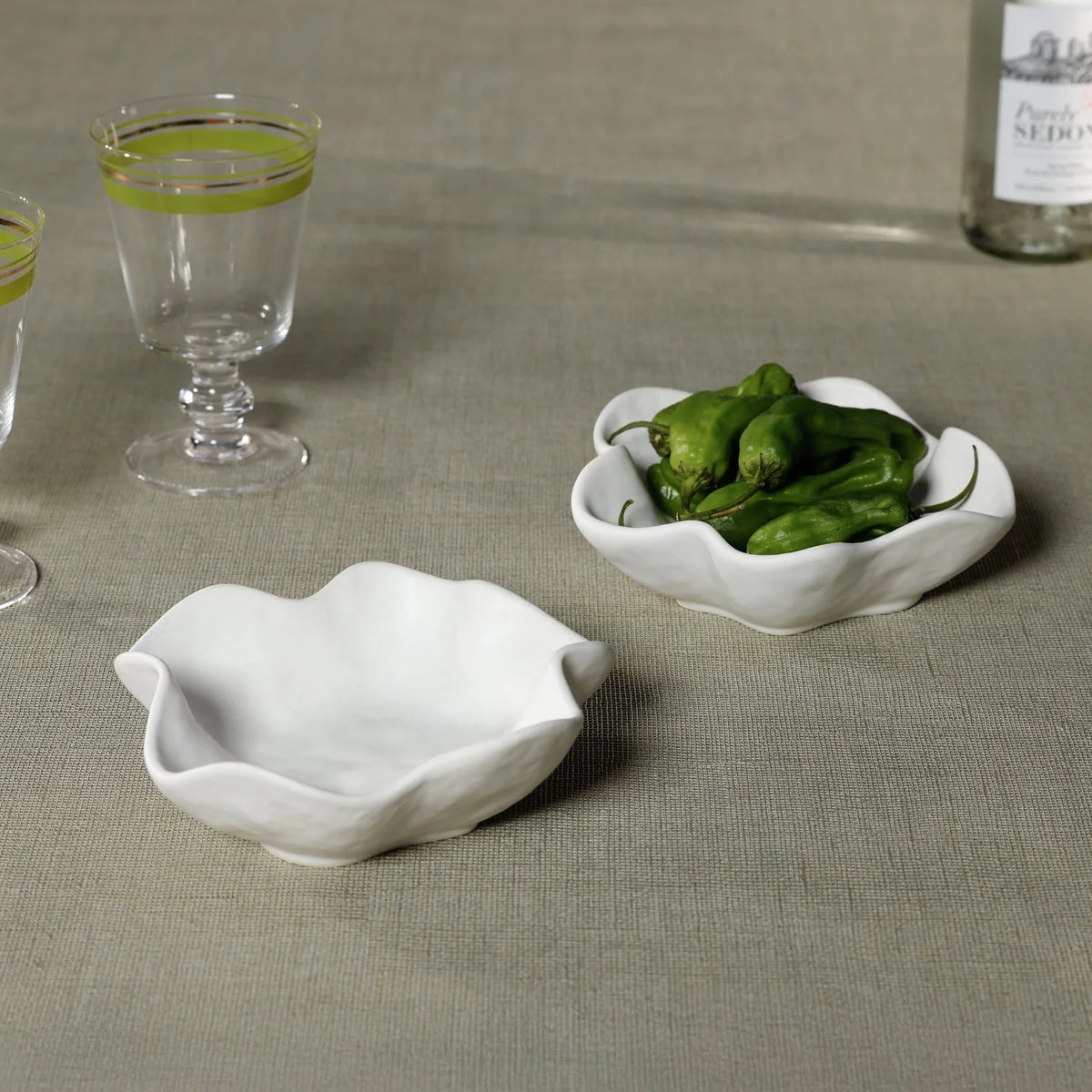 Two Zodax Alto Ceramic Condiment Bowls with wavy edges sit on a beige surface—one empty, one with green peppers. Nearby are two clear glasses with green stripes and a partially visible bottle.