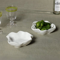 Two Zodax Alto Ceramic Condiment Bowls with wavy edges sit on a beige surface—one empty, one with green peppers. Nearby are two clear glasses with green stripes and a partially visible bottle.