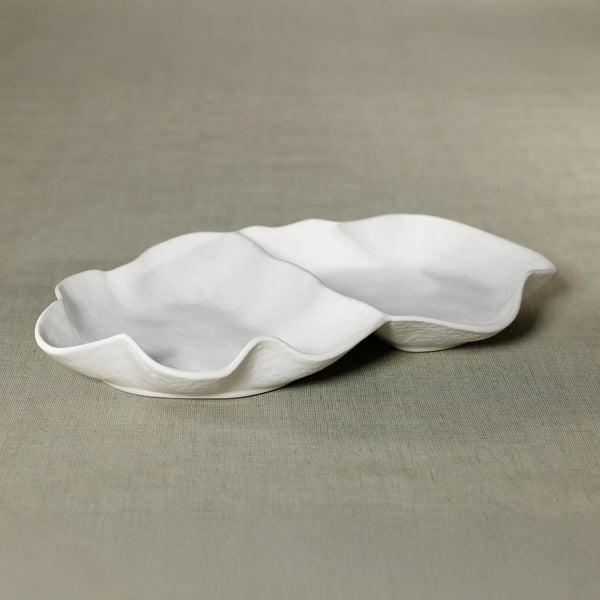 The Alto Two Section Ceramic Serving Bowl by Zodax, featuring a matte white petal-like design with wavy edges, rests on a muted green textured surface.