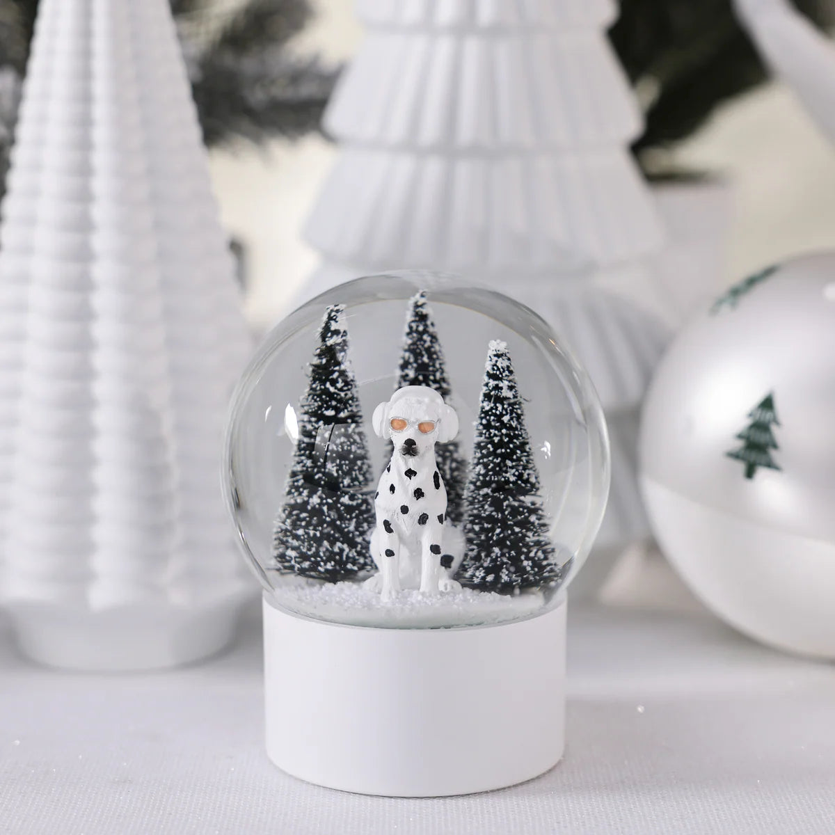 A Zodax Dalmation Snow Globe featuring a dog wearing orange sunglasses, standing in front of two pine trees on a snowy base.