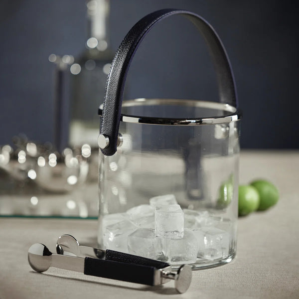 The Zodax Laguna Ice Bucket features a glass design with a black handle, filled with ice cubes. Metal tongs with black grips rest in front, while limes, a glass bottle, and barware accent the scene. Ideal as a wine or champagne chiller.