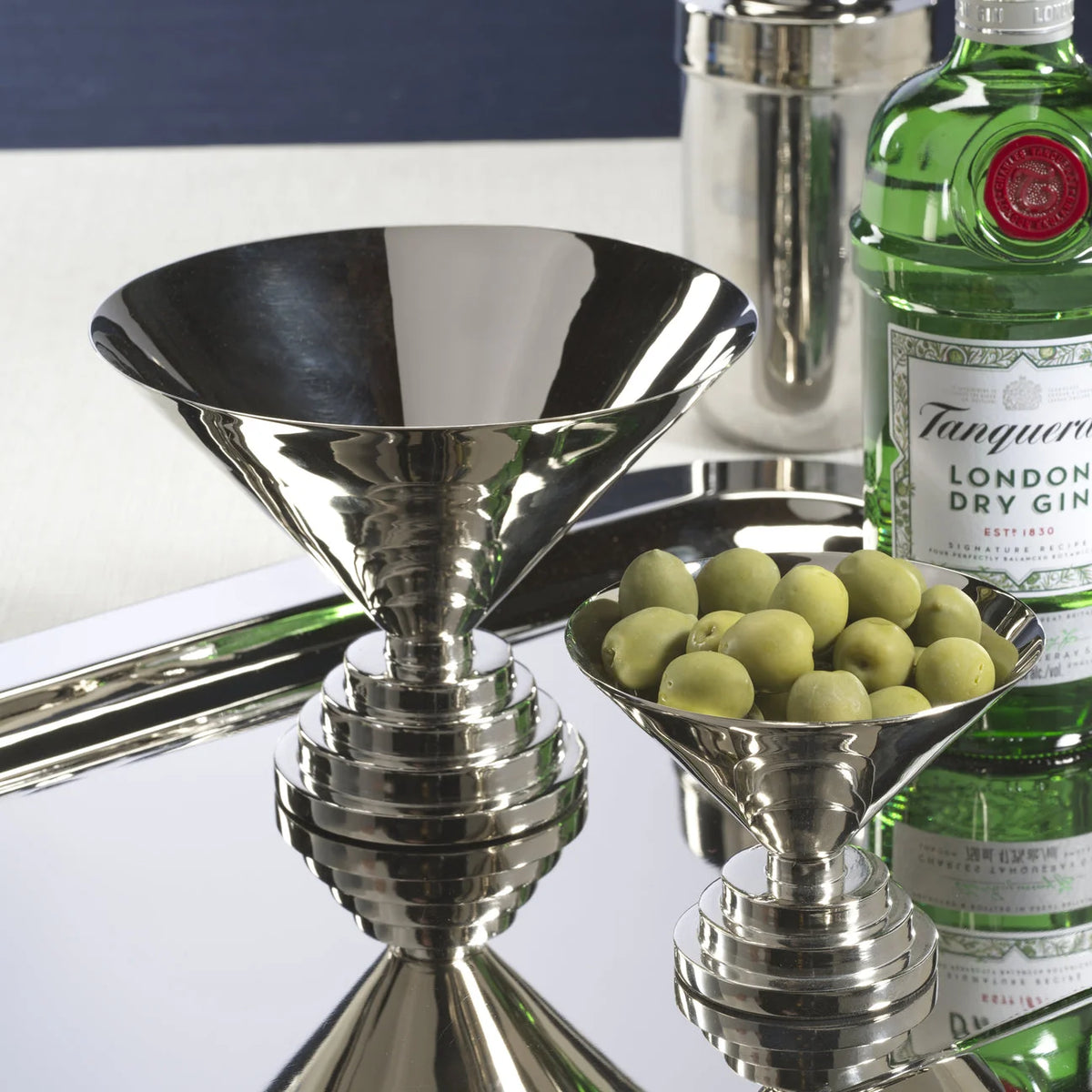 A silver martini glass, the Zodax Malcolm Condiment Bowl in polished nickel filled with green olives, a bottle of Tanqueray gin, and a cocktail shaker are arranged on a reflective tray in striking Art Deco style.