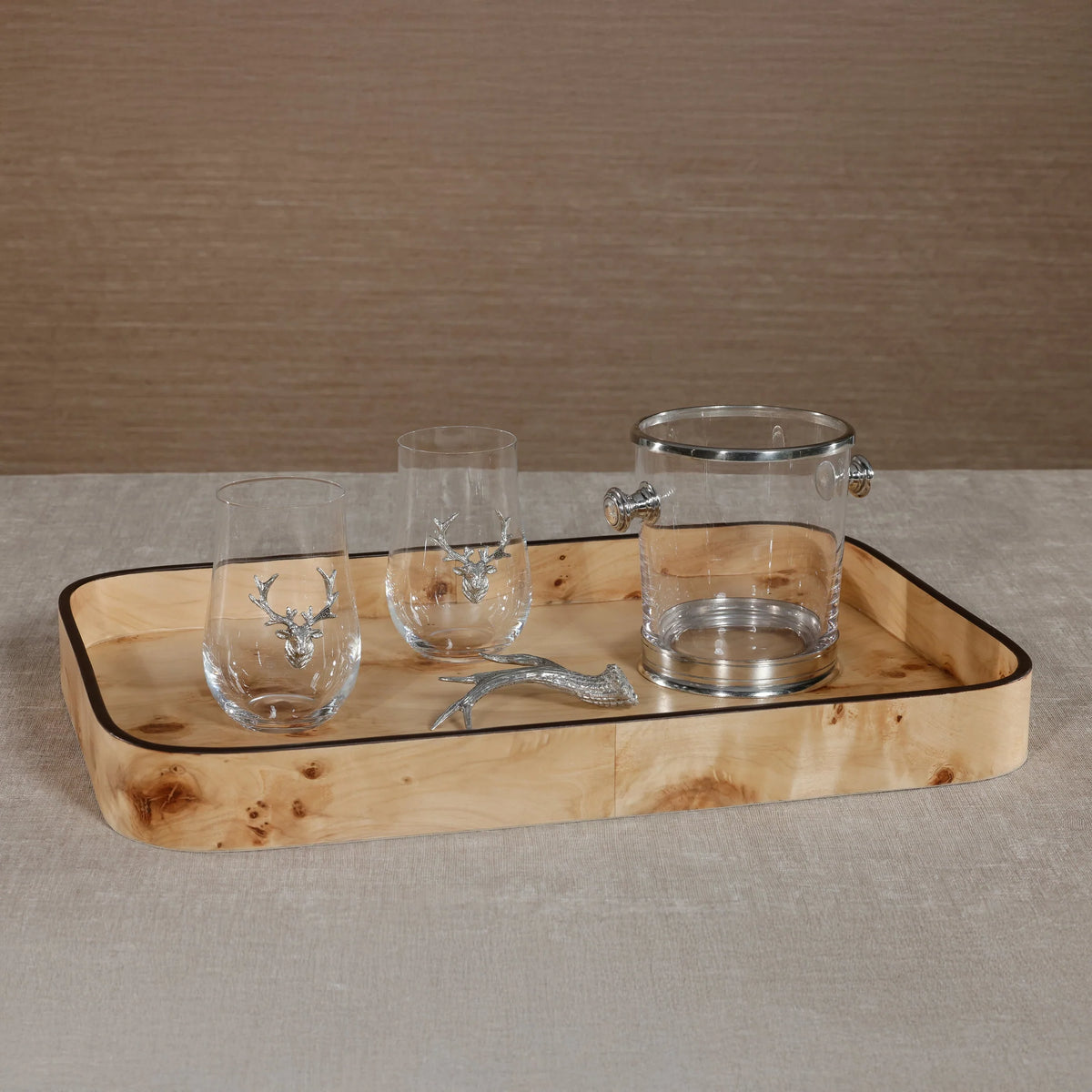 The Zodax Valari Rectangle Serving Tray in light burl veneer holds two stemless glasses with silver deer accents, a clear ice bucket with a silver lid, and a deer antler-shaped silver bottle opener on a beige surface.