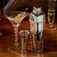 The Juliska Graham Cocktail Shaker is elegantly arranged on a polished wooden surface beside a martini garnished with green olives, silver stirring sticks, and a glass of champagne.