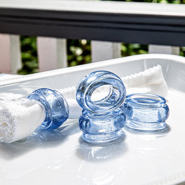 Four Provence Napkin Rings by Juliska are displayed on a white tray, one holding a folded white napkin. Set outdoors with greenery, the rings add vibrant color and style to the table. Sold as a set of 4.
