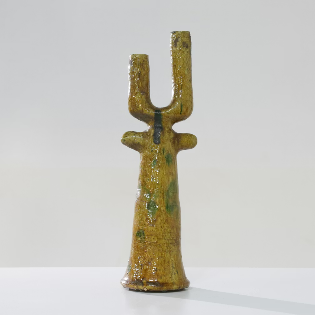 The Govad Candleholder 20″ by Schwung Design, a yellow ceramic piece with a textured, glossy finish and an elongated form featuring two upward prongs and two small side protrusions reminiscent of Moroccan-inspired handmade candlesticks, stands on a white surface against a plain light background.