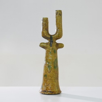 The Govad Candleholder 20″ by Schwung Design, a yellow ceramic piece with a textured, glossy finish and an elongated form featuring two upward prongs and two small side protrusions reminiscent of Moroccan-inspired handmade candlesticks, stands on a white surface against a plain light background.