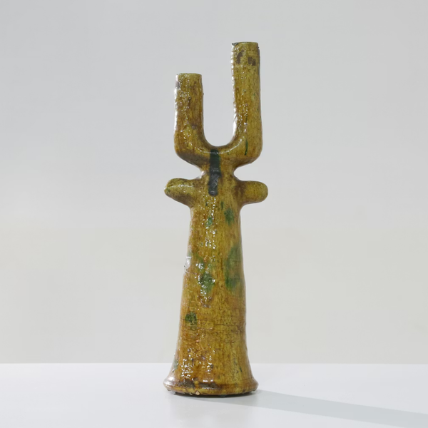 The Govad Candleholder 20″ by Schwung Design, a yellow ceramic piece with a textured, glossy finish and an elongated form featuring two upward prongs and two small side protrusions reminiscent of Moroccan-inspired handmade candlesticks, stands on a white surface against a plain light background.