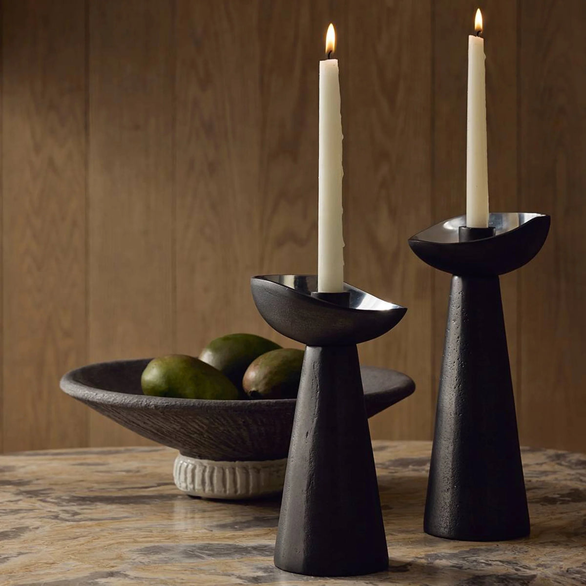 Two tall Argemone Candleholder Set of 2 - Black by Arteriors with white candles stand on a marble surface. In the background, a dark bowl with green fruits sits before a wooden panel wall.