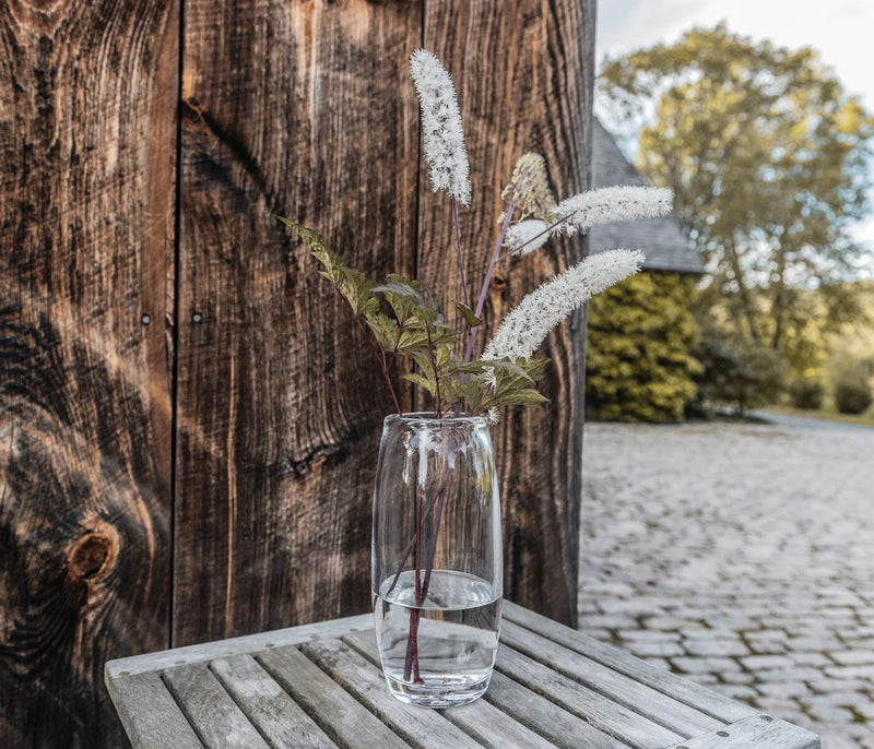 A Simon Pearce Addison Vase holds three white fluffy flowers with green leaves, displayed on a wooden table outdoors near a weathered wall, with trees and cobblestones providing a charming background.