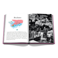 An open Assouline Amalfi Coast Book reveals a watercolor map marked "Amalfi Coast" and "Positano" on the left page, with a black-and-white hillside town photo featuring a person on a terrace on the right.