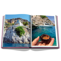 The Amalfi Coast Book by Assouline displays two photos: a stone building perched on a seaside cliff, and a hand holding a sea urchin above clear blue water with a boat in the background.