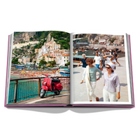 An open Assouline Amalfi Coast Book displays a red scooter by a seaside railing in a vibrant coastal town on the left page, with stylishly dressed people strolling along the beach on the right.