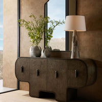 A modern sideboard with rounded edges displays the elegant Arteriors Amal Grey Glazed Vases Set of 2, filled with green branches, alongside a stacked stone lamp. A large mirror reflects the desert landscape visible through a tall window, while neutral-toned walls create a serene backdrop.