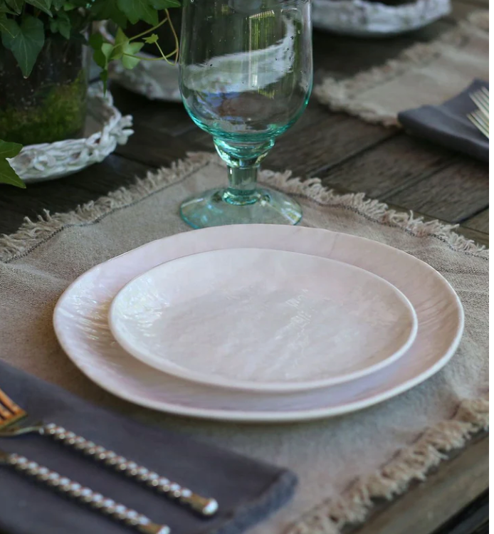 A close-up of a rustic table set with Relish Weave Melamine Dinnerware in white, a glass goblet, silver cutlery on a dark napkin, and a beige fringed placemat on wood. Greenery appears in the background.