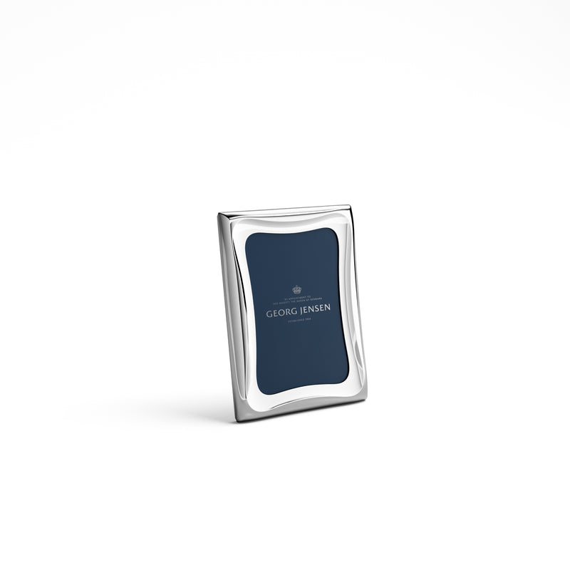 The Georg Jensen Cobra Stainless Picture Frame features sleek silver stainless steel with smooth, wavy edges. It stands upright on a white background with a navy blue insert reading "Georg Jensen" in white text.