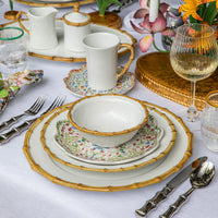 Juliska’s Bamboo Dinnerware creates an elegant table setting with its white and floral-patterned design, complemented by gold-rimmed edges. The arrangement features glassware, silver cutlery, decorative flowers on a white tablecloth, a gold serving platter, and a wine glass to complete the look.