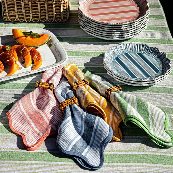 A table set with Juliska's Cabana Stripe Napkin Set of 4 - Multi Set, striped plates, napkins in rings, and a melon prosciutto platter atop a striped linen tablecloth and a woven basket to complete the vibrant look.