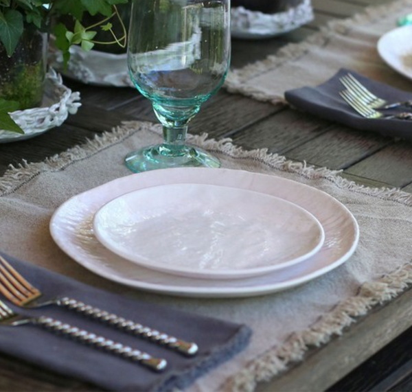 A close-up of a rustic table set with Relish's Weave Melamine Dinnerware in white, displayed on a beige placemat atop a wooden surface, with cutlery on a gray napkin, clear water glass, and plants in the background.