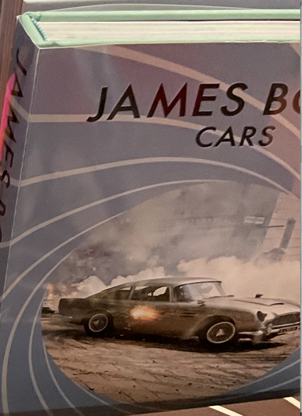 A close-up of the Assouline book "James Bond Cars," showcasing the cover with a classic car driving through smoke and flames, set against swirling background lines.
