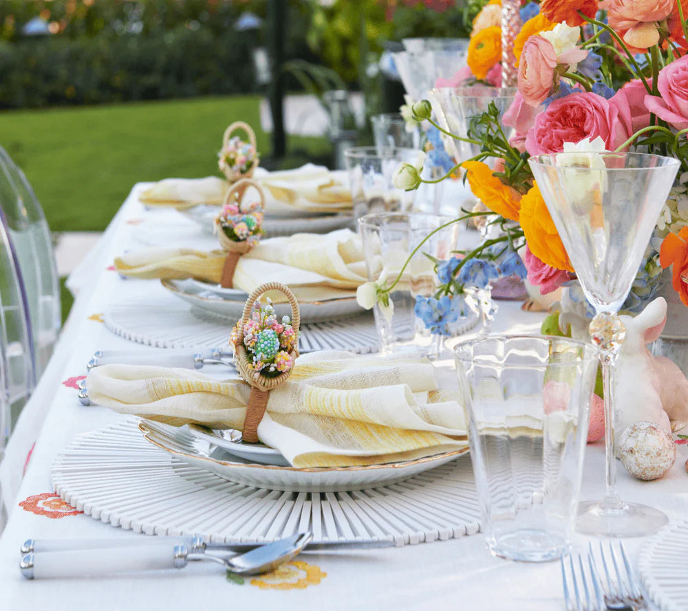An outdoor table features white plates, Kim Seybert Broad Stripe Napkin Set of 4 in yellow & white, wicker napkin rings with pastel candies, clear glasses, and bold floral centerpieces with pink, orange, and yellow blooms.