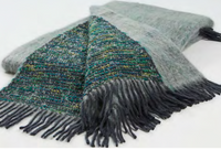 A folded Coco Throw by Mantas Ezcaray with gray, blue, and green tones, featuring a textured pattern and long fringe along the edges, set against a white background. The double-sided design offers versatility and style, making this Mohair throw a cozy yet elegant accent for any space.