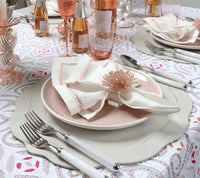 A refined table setting features white and pink plates on Kim Seybert’s Tailored Placemat Set of 4 in Ivory, paired with white napkins with pink trim, beaded napkin rings, silverware, glassware, and rose-hued drinks on a floral tablecloth.
