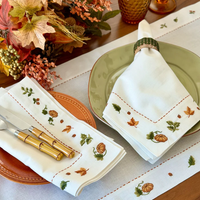 Create a cozy Thanksgiving atmosphere with an elegant fall table setting featuring green and orange plates, gold-handled cutlery, and the Bodrum Autumn Medley Napkin Set of 4. These white napkins are adorned with pumpkins and autumn leaves, adding a festive touch to your gathering. The arrangement is completed by a striking floral centerpiece framed by seasonal autumn foliage, inviting guests to celebrate in style.