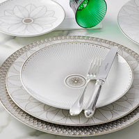 A set table featuring patterned white and platinum plates from the Malmaison Imperiale Porcelain Dinnerware collection by Christofle, with a knife and fork resting on the top plate and a green glass cup in the background atop a marble surface—an elegant showcase of Christofle’s luxury tableware.