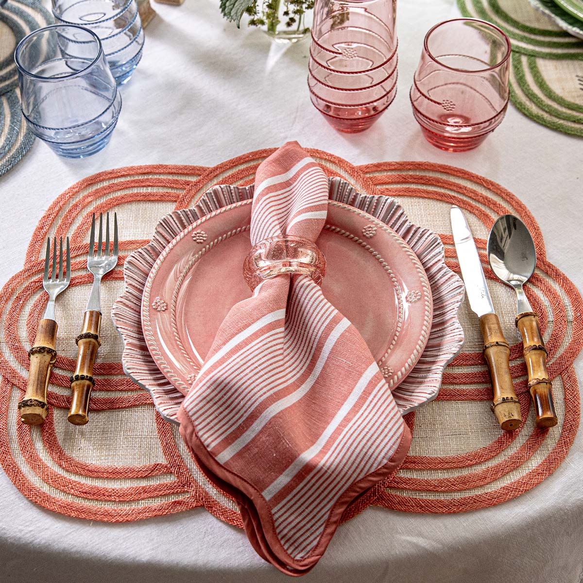 A lively tabletop features a pink plate, Juliska’s Cabana Stripe Napkin Set of 4 in Coral, bamboo-handled cutlery, pink and blue glasses, coral placemats, and a white tablecloth. Green placemats add color to the scene.