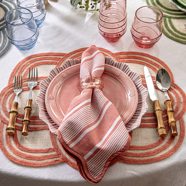 A lively tabletop features a pink plate, Juliska’s Cabana Stripe Napkin Set of 4 in Coral, bamboo-handled cutlery, pink and blue glasses, coral placemats, and a white tablecloth. Green placemats add color to the scene.